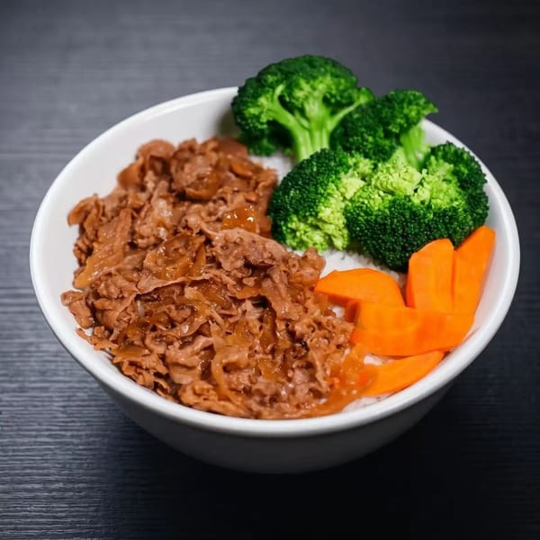 A white bowl of gyudon with sliced beef, broccoli florets, and carrot slices on a dark surface.
