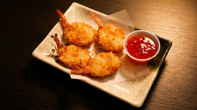 Four crispy coconut shrimp with tails on, served with a side of red sweet chili dipping sauce.