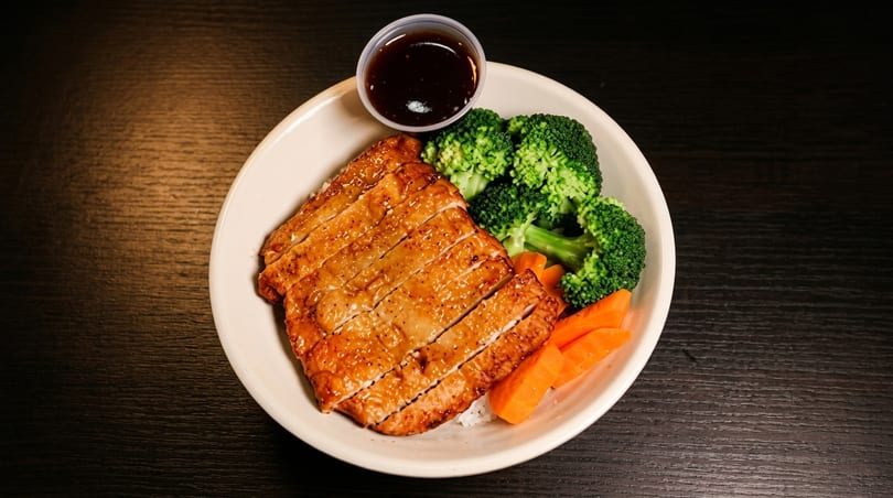 Sliced grilled chicken with broccoli, carrots, and a side of sauce in a white bowl.