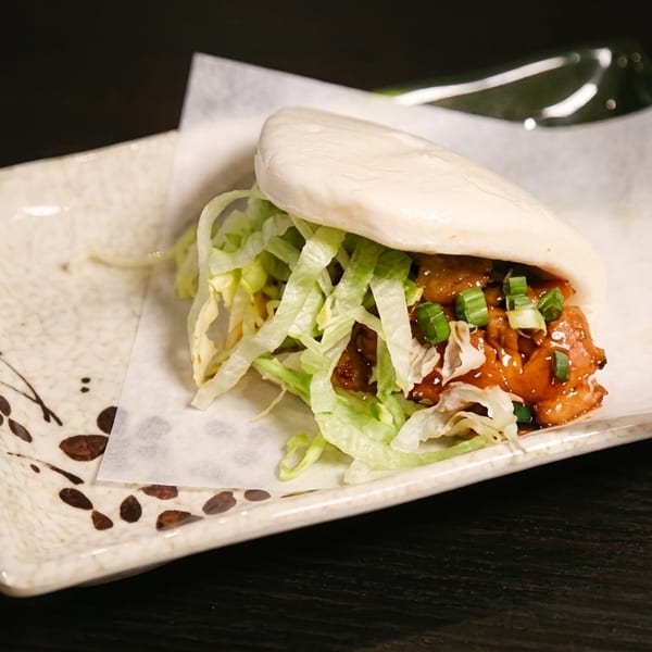 A fluffy bao bun filled with glazed chicken, shredded lettuce, and green onions on a decorative plate.