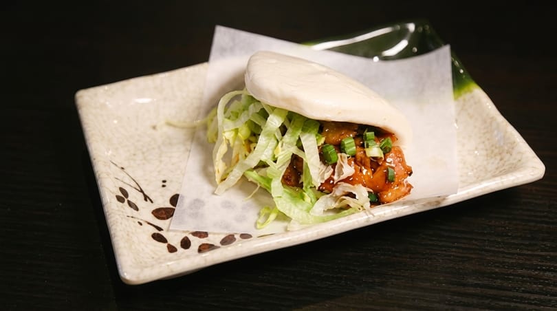 A fluffy bao bun filled with glazed chicken, shredded lettuce, and green onions on a decorative plate.