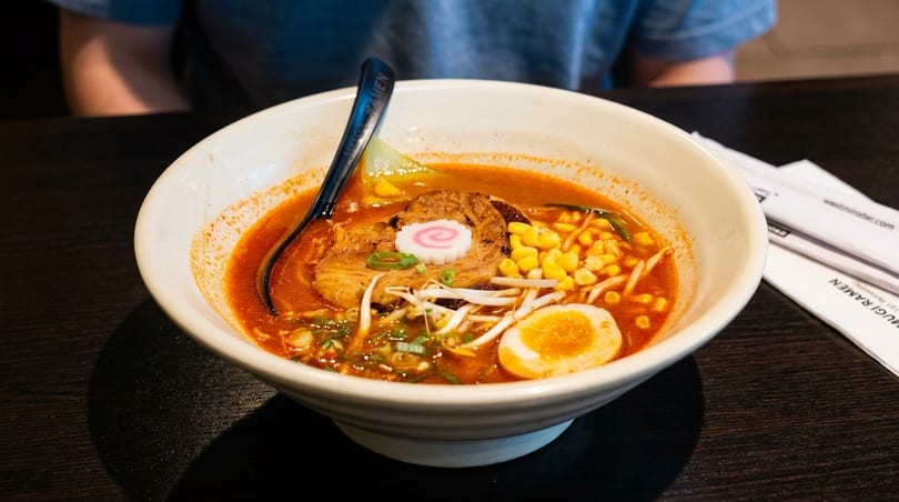 Spicy ramen with pork, corn, egg, and fish cake in a white bowl on a dark table.